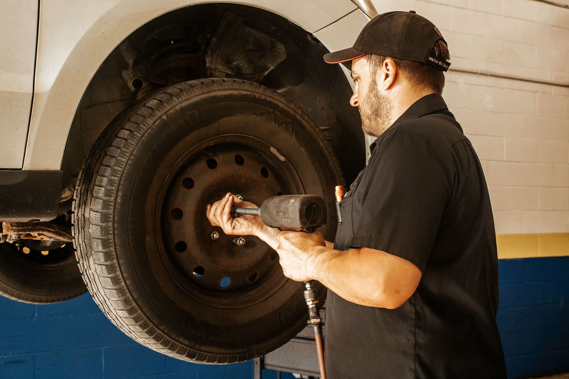 Mechanic in black shirt and cap uses a wrench to remove a tire from a white car in a garage.