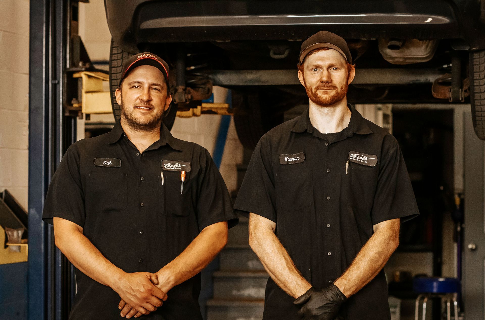 Two mechanics in black shirts and caps, posing in a car repair shop. One has black gloves.