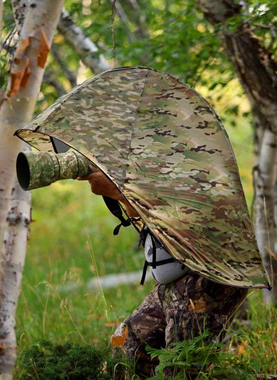 Photographer hidden under camouflage cover, holding camera with a long lens in a forest setting.