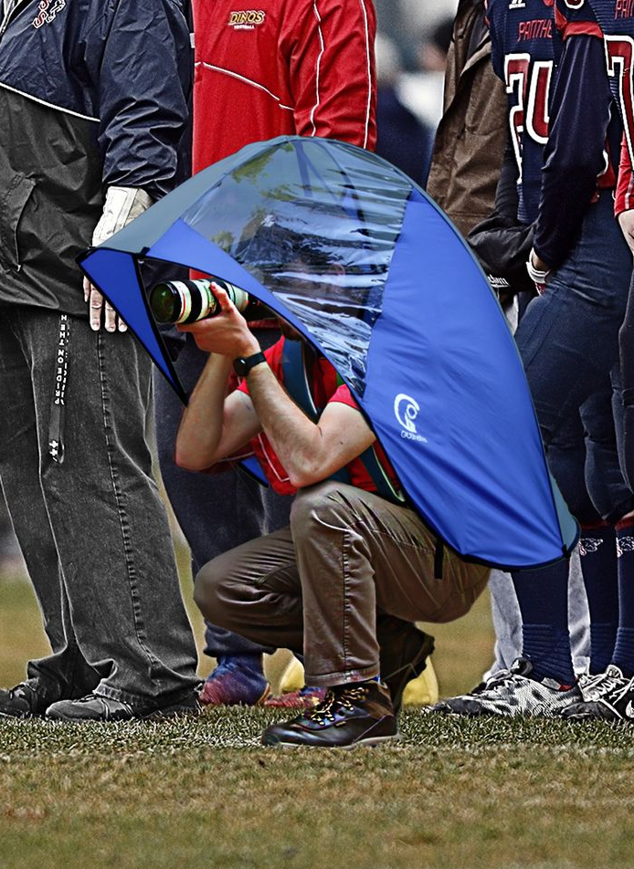 Photographer crouches under a blue umbrella, taking photos at an outdoor sports event.