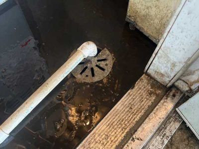 Flooded floor with drain and PVC pipe. Dirty water surrounds a drain in a tiled room next to a white cabinet.