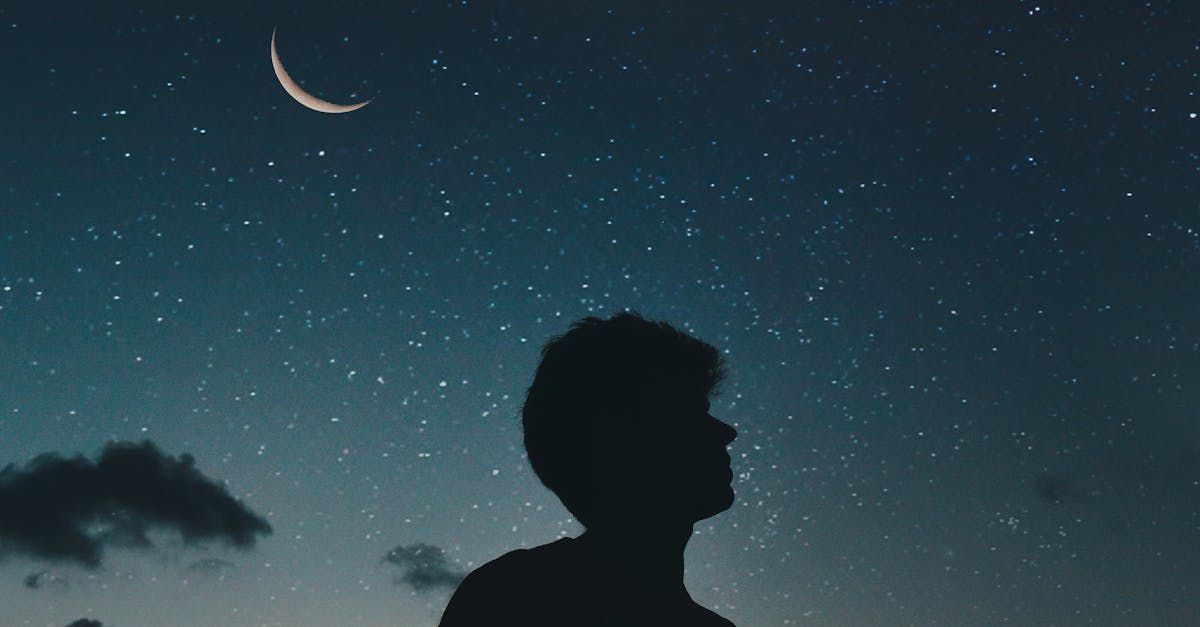 A silhouette of a man looking at a crescent moon in the night sky.
