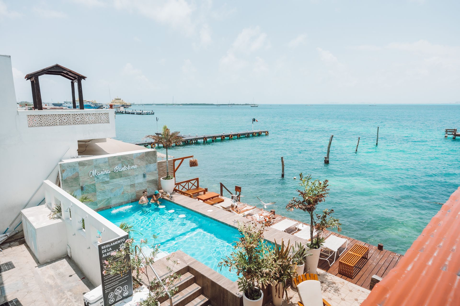 A pool with people next to the turquoise ocean, a pier and blue sky.