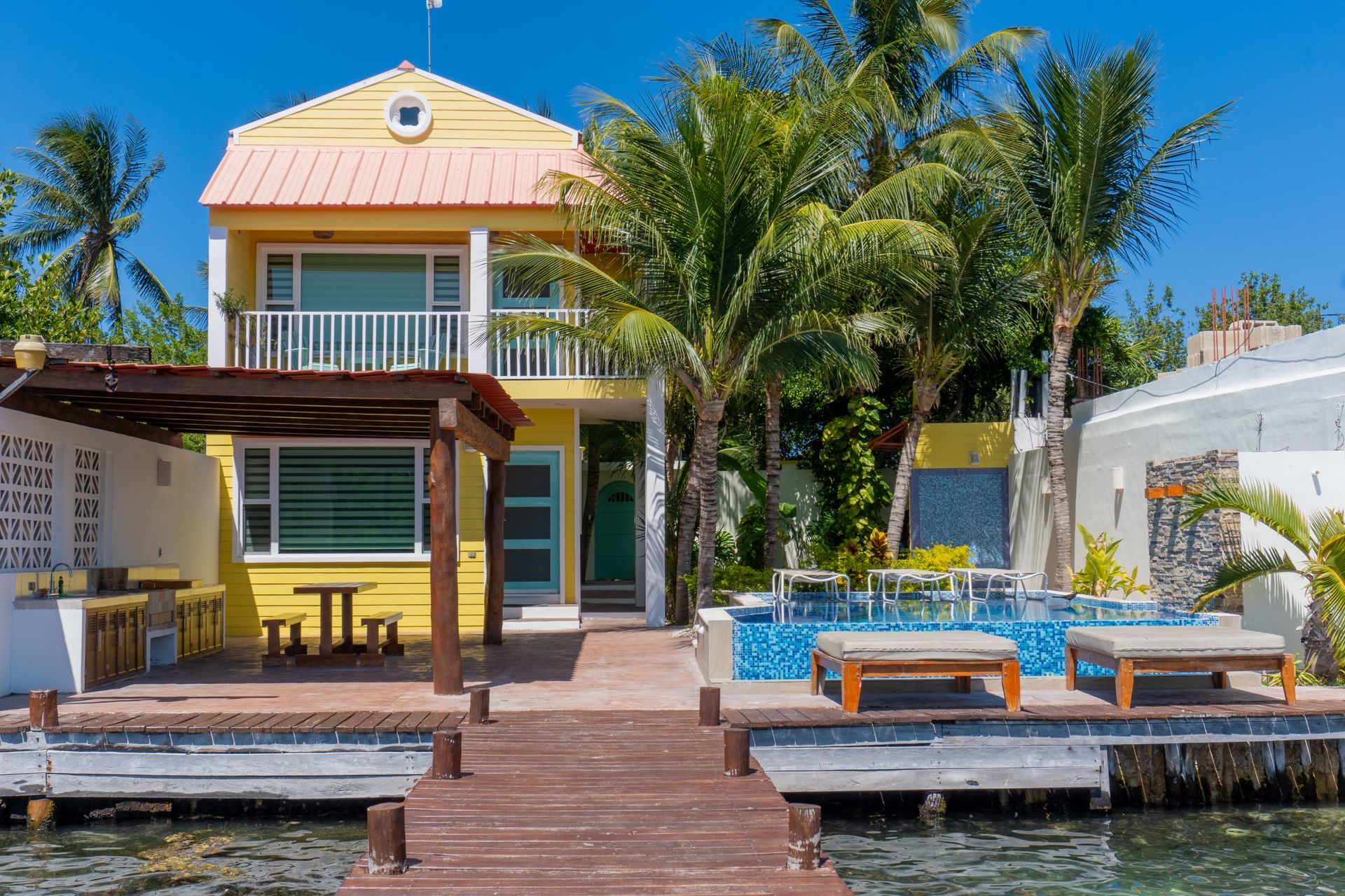 Yellow tropical house with dock, small pool, and palm trees under a bright blue sky.