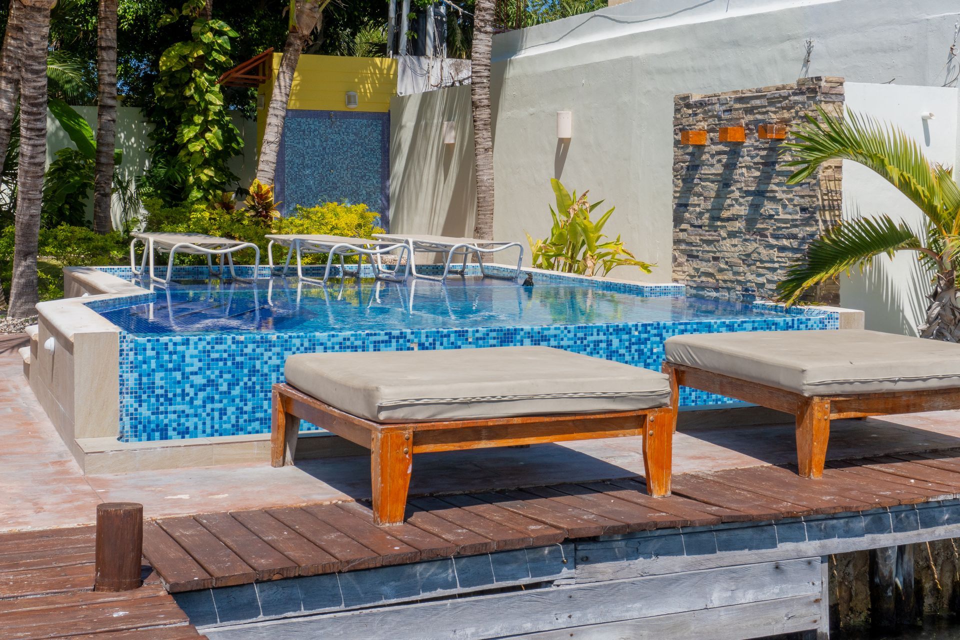 Pool with blue tile, wooden deck, lounge chairs. Palm trees, blue mosaic wall.