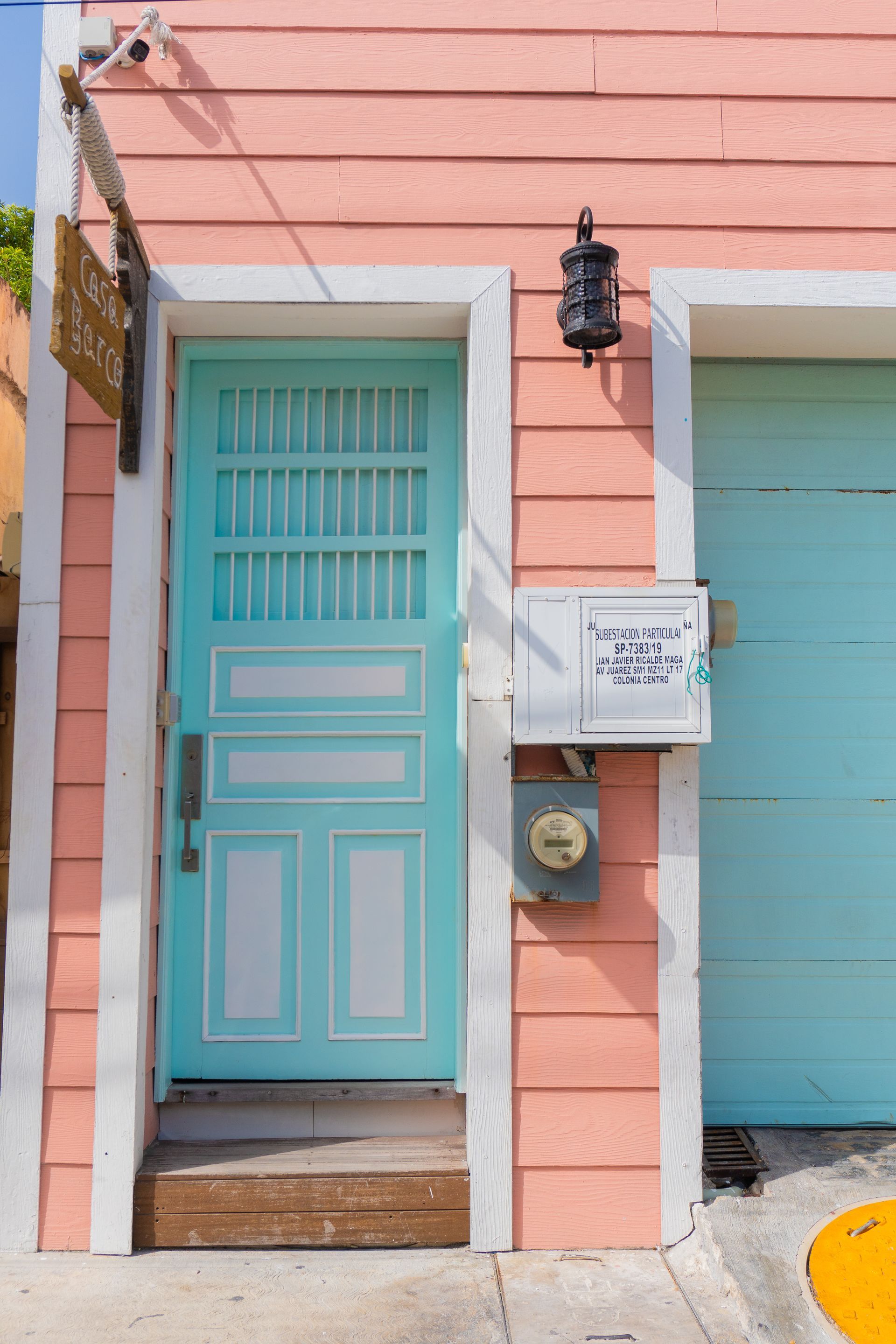 Pink and blue exterior: aqua door, garage, and trim against a pink clapboard wall. A sign hangs beside the door.