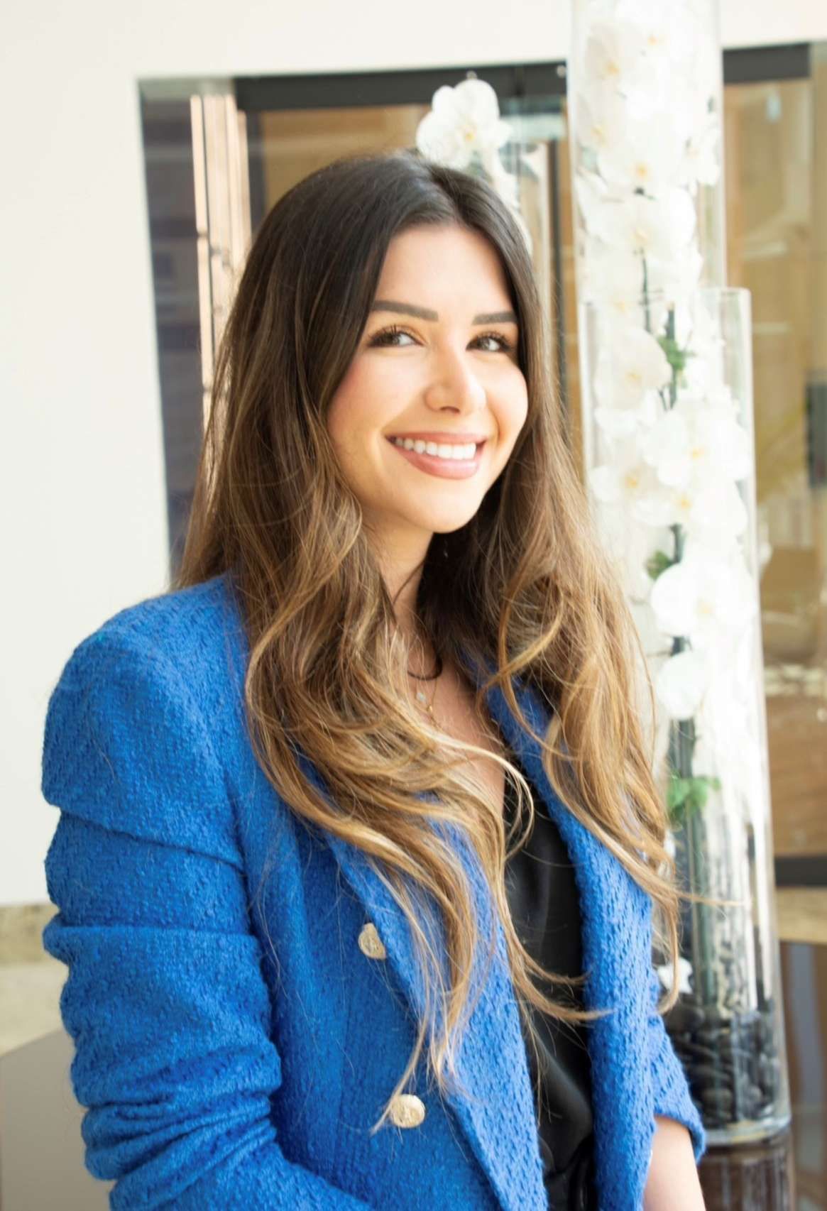 Woman with long brown hair smiles, wearing a blue blazer, standing near white flowers.