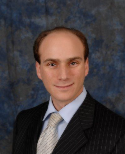 Studio portrait of a person in a dark pinstripe suit and light tie against a blue-gray backdrop