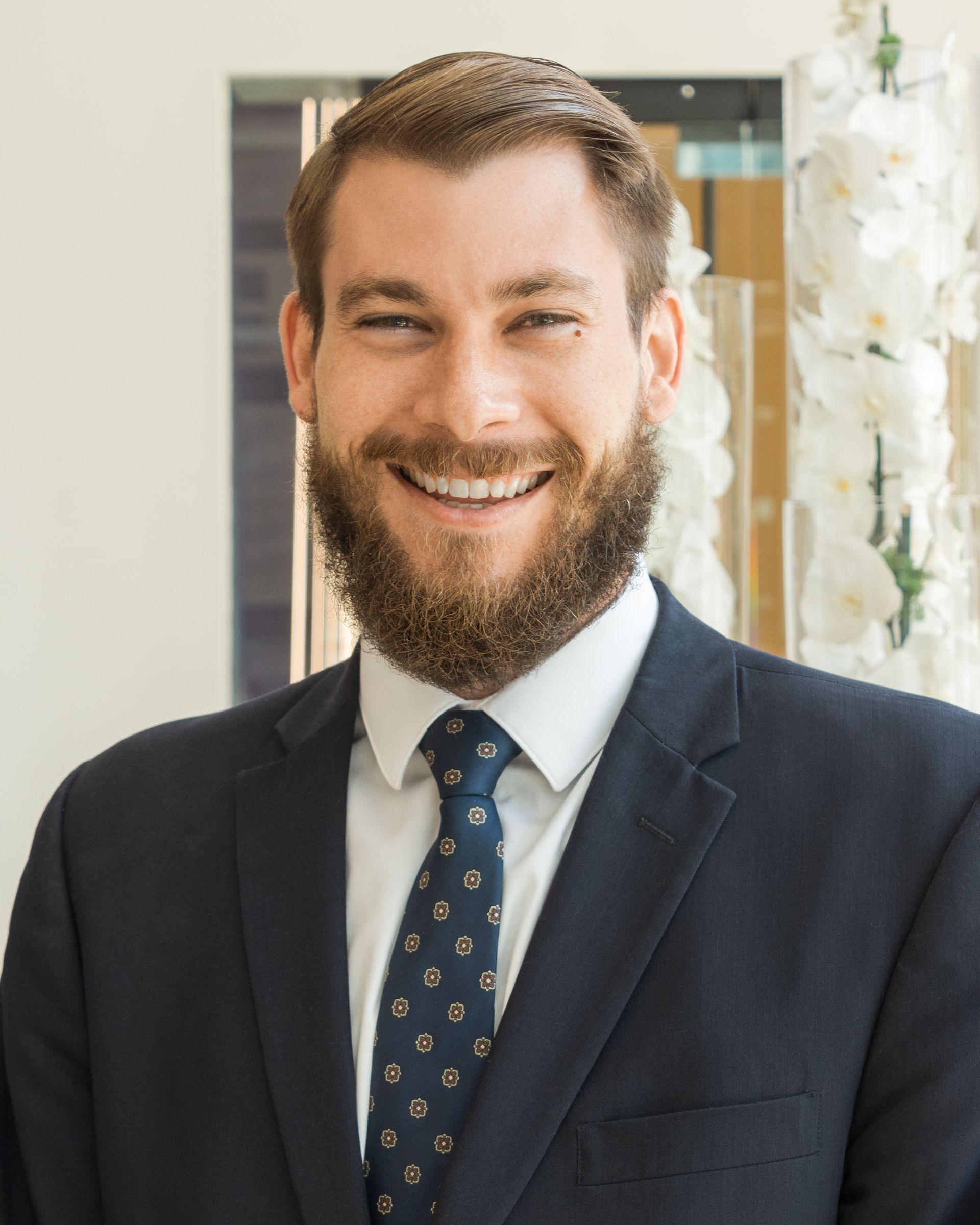 Man in a suit smiles at the camera. He has a beard, tie, and is indoors near flowers.
