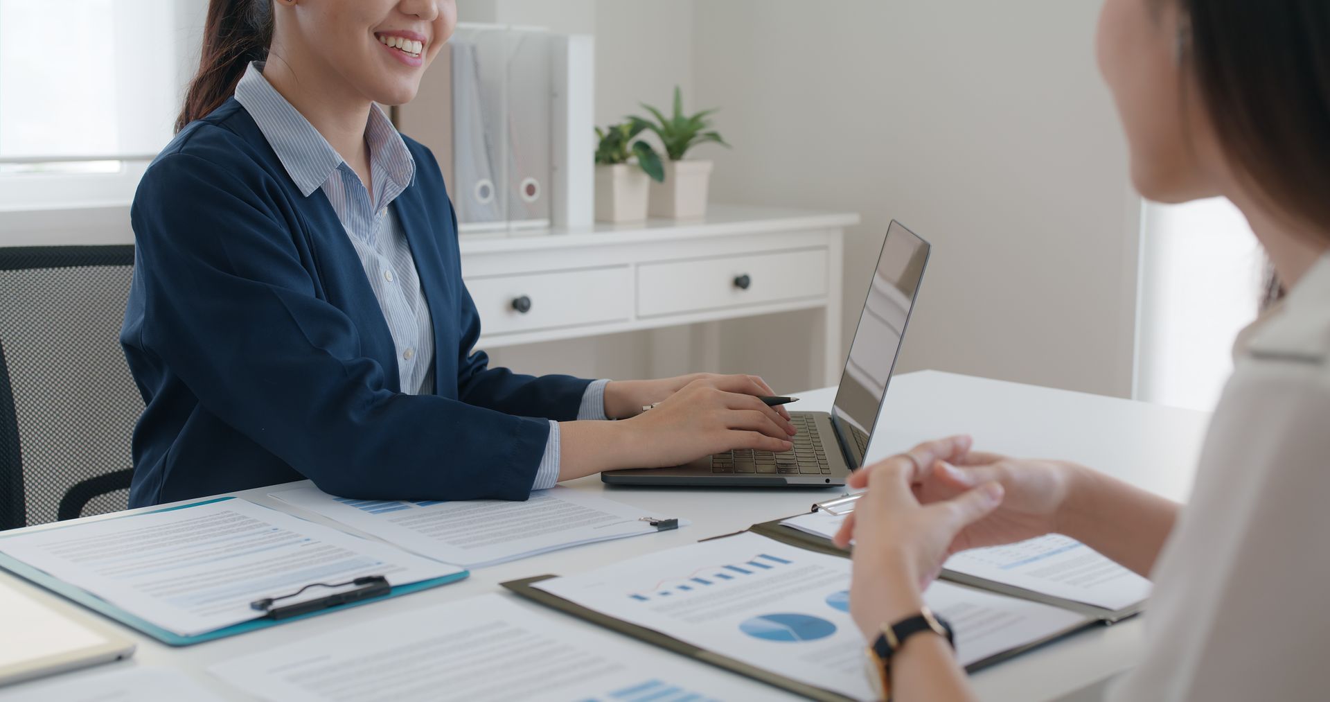 Woman in blue blazer types on laptop, smiling at a person seated across a desk with documents.