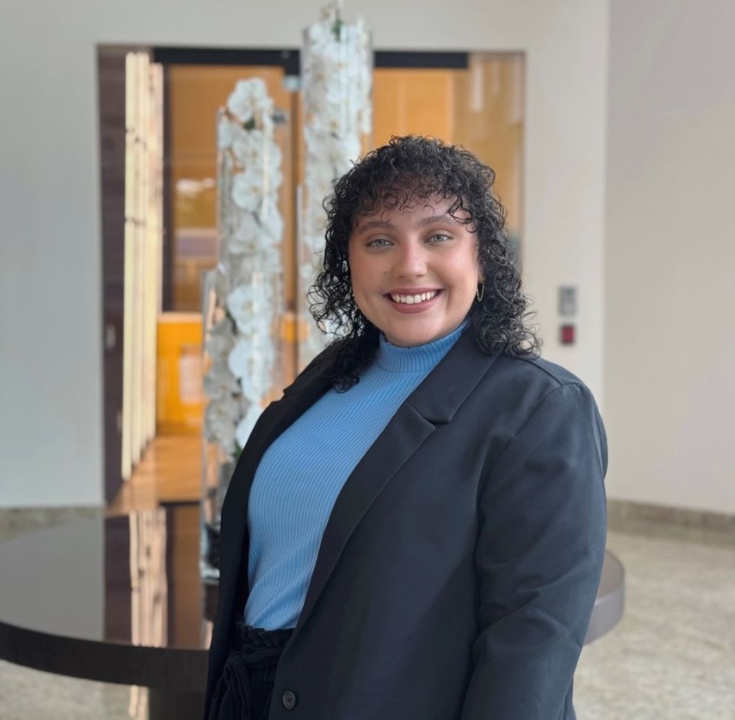 Woman with curly hair smiles, wearing a blue top and black blazer, standing in a lobby.