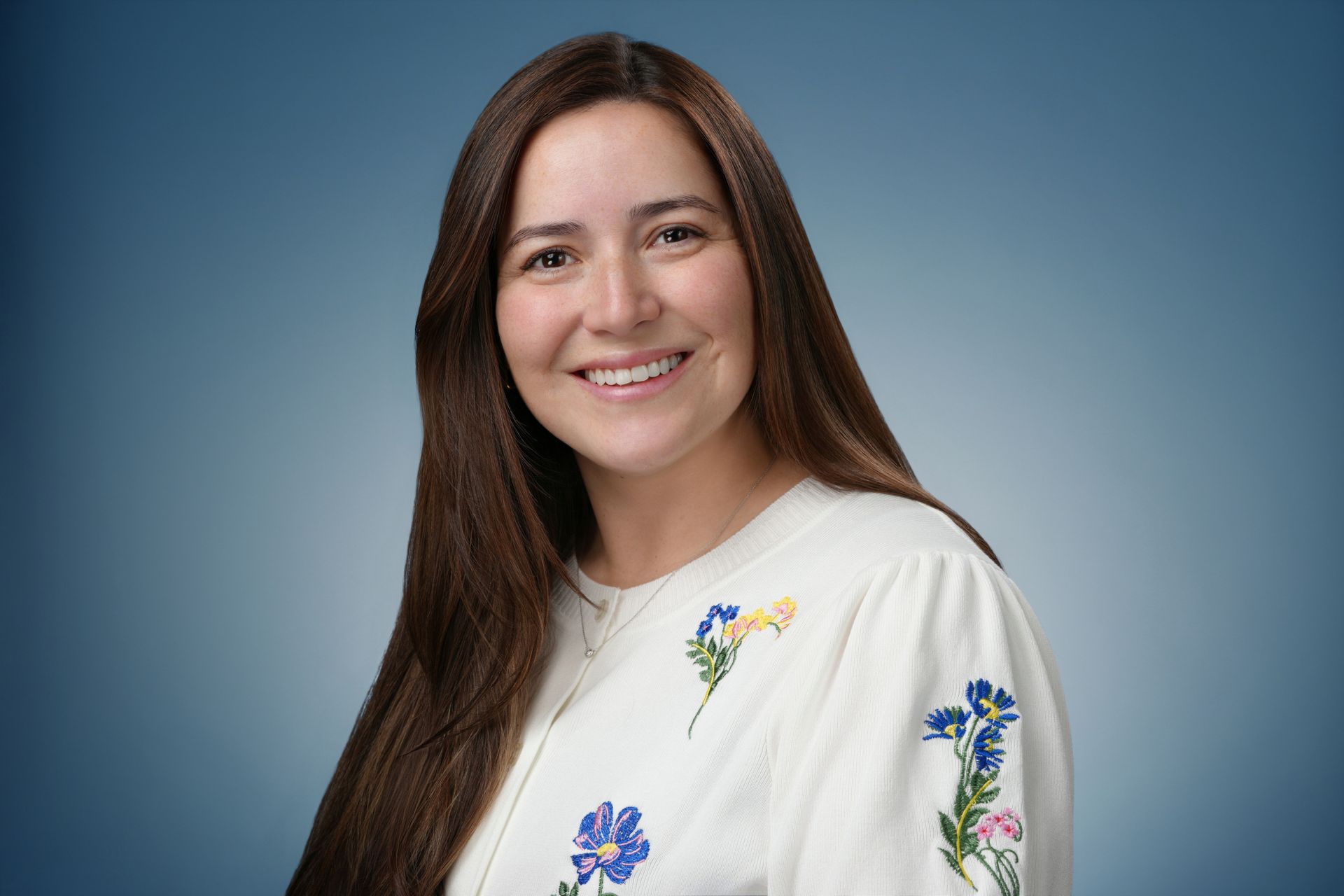 Woman with long dark hair, smiling. Wearing a white sweater with black bows.