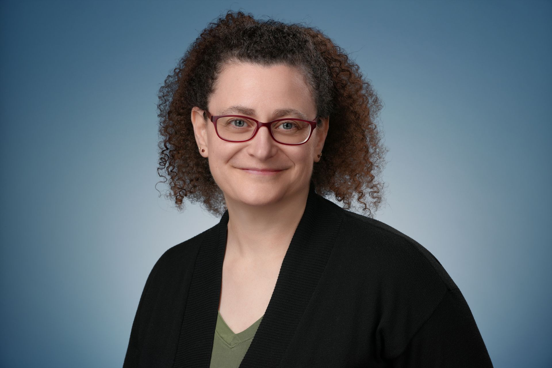 Woman with curly hair and glasses smiles at the camera, wearing a black vest against a blurred blue background.