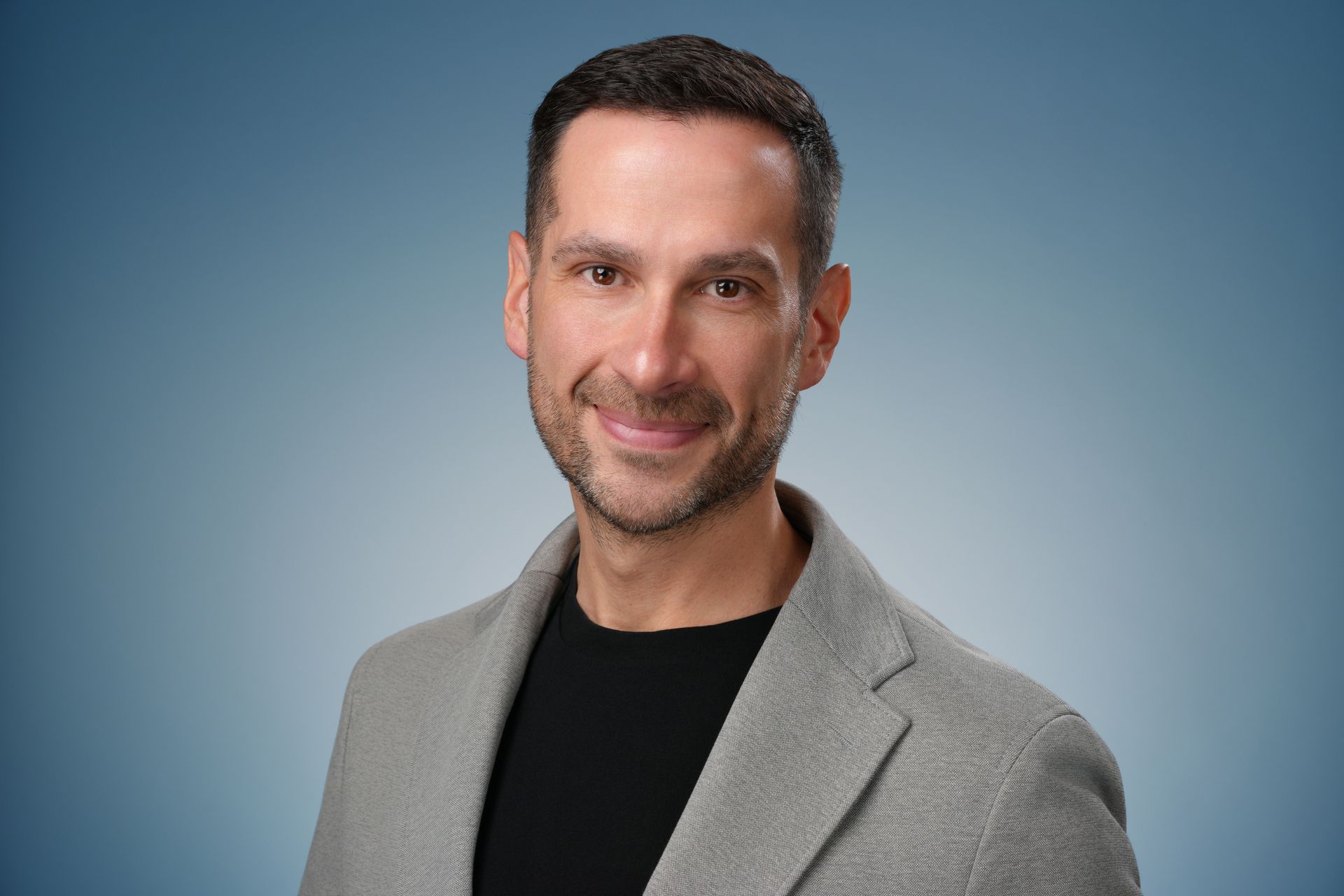 Man with brown hair and beard smiles broadly in front of a blue backdrop. He wears a black shirt and a jacket.