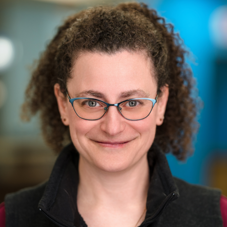 Woman with curly brown hair wearing glasses, smiling, and looking at the camera.