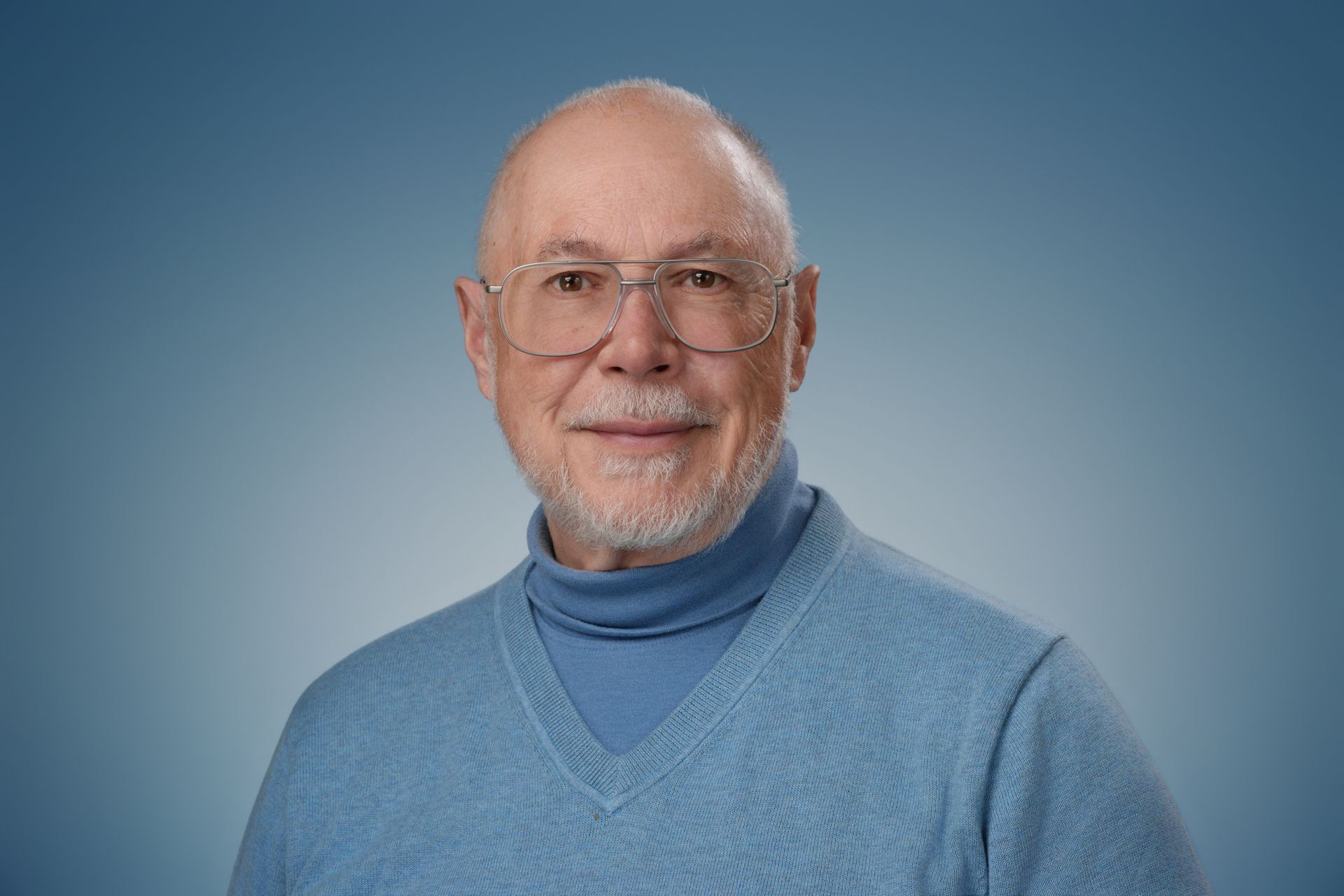 Bald man with glasses and gray beard smiles, wearing a gray and black zipped jacket; blurred blue background.