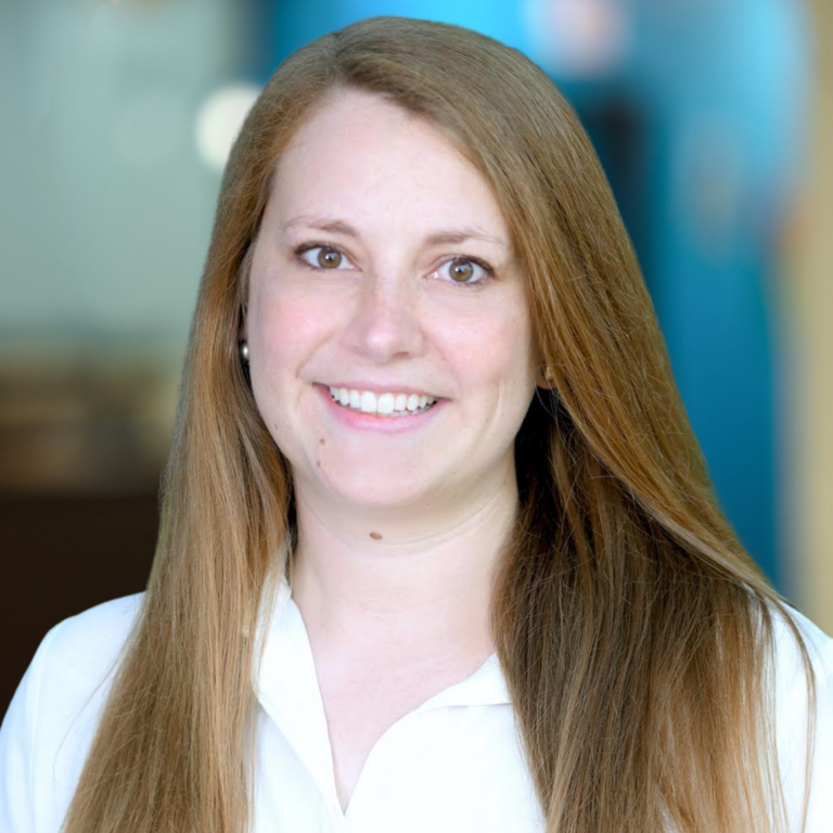 Woman with long brown hair smiles, wearing a white shirt; blurred background.