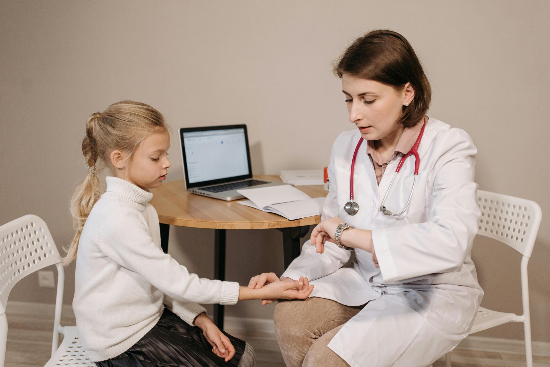 Doctor checking a child's pulse with stethoscope buring a check-up. They sit at a table with a laptop.