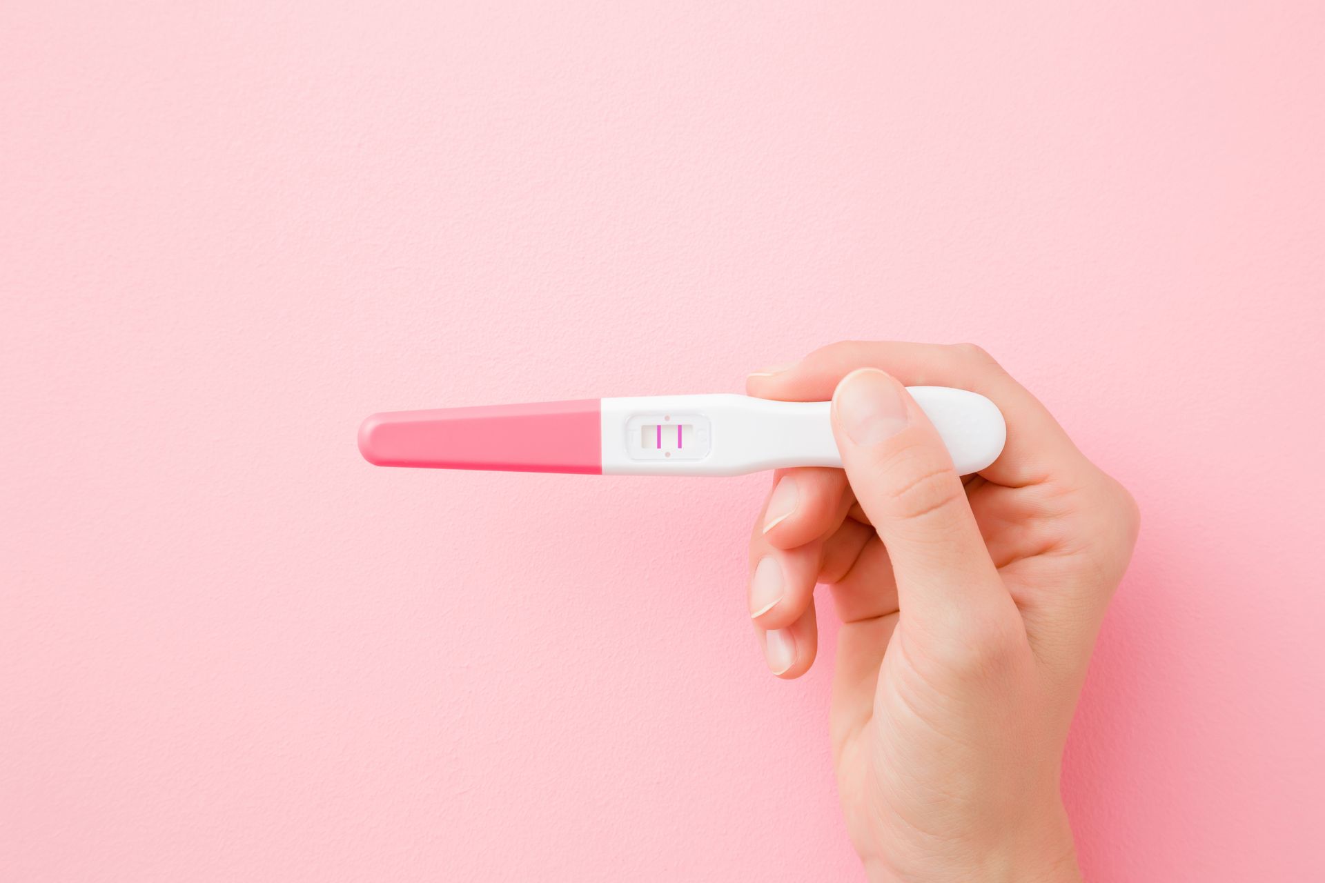 Hand holding a positive pregnancy test showing two lines against a soft pink background.