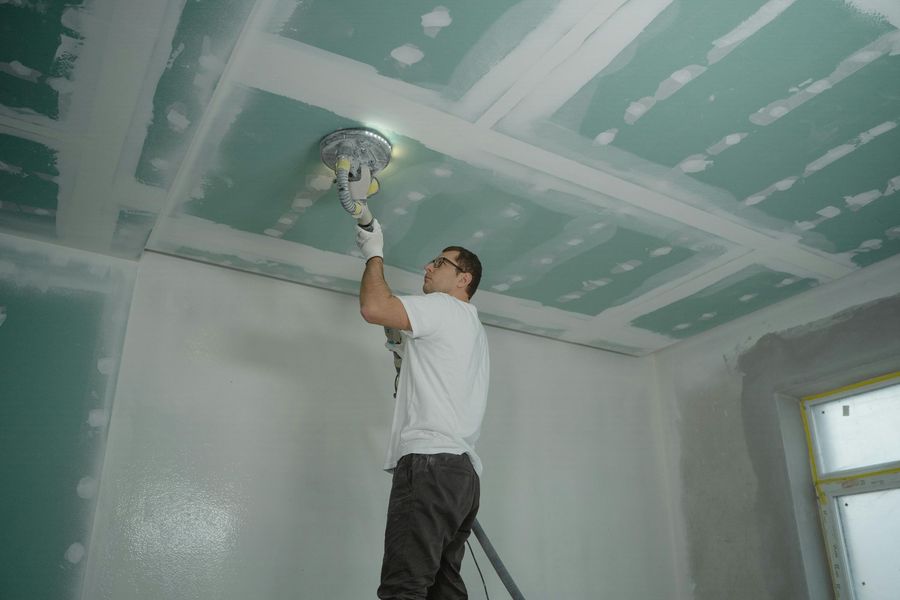 Man sanding drywall ceiling with sander, wearing gloves, in a room with partially finished walls.