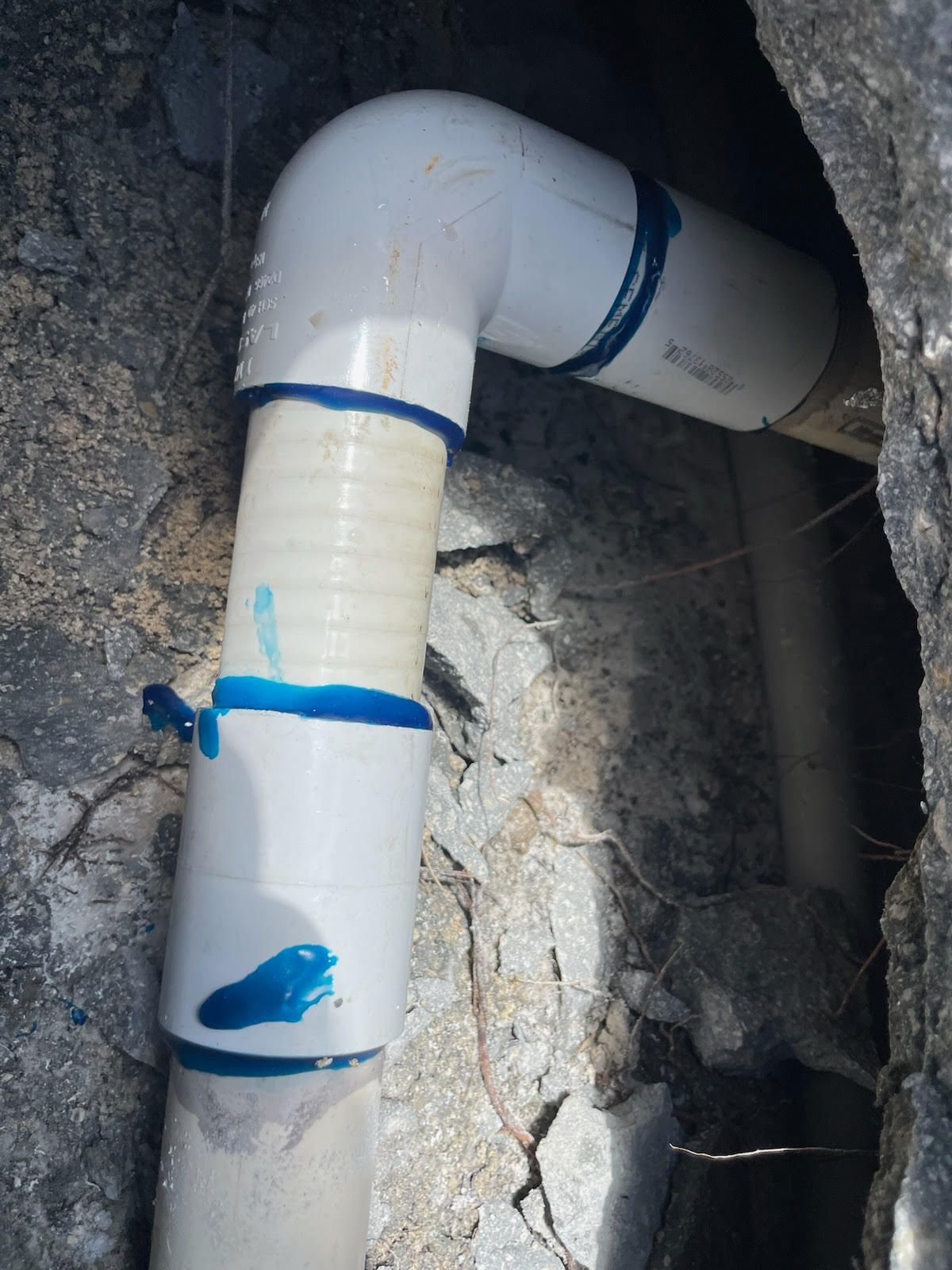 White PVC pipe with blue tape bends through a rough hole in a concrete or rock wall.