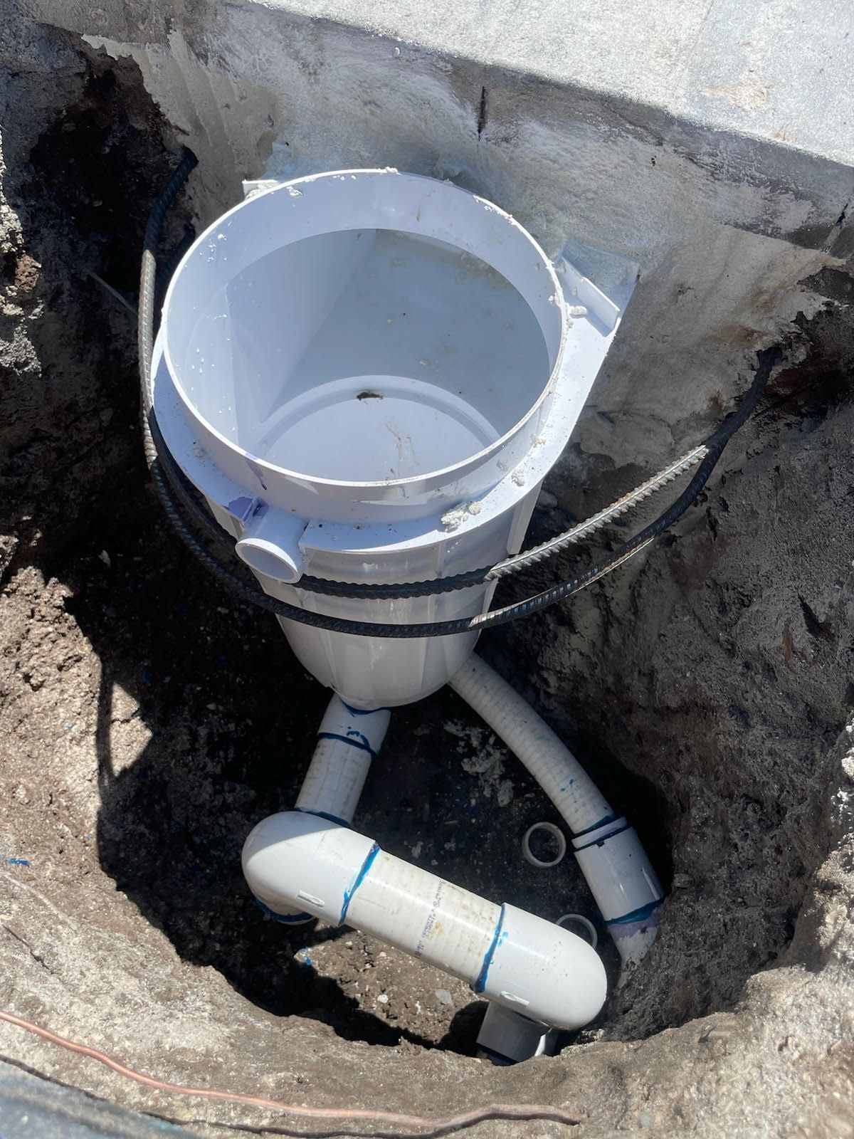 White PVC pipe fitting set in a dirt excavation beside a concrete wall.