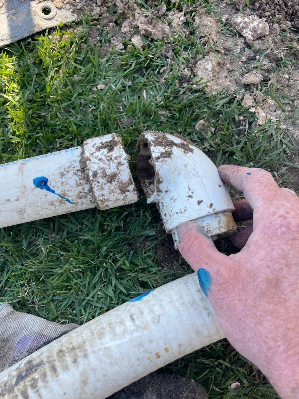 Hand holding a broken white PVC pipe elbow over grass, with another pipe section nearby.