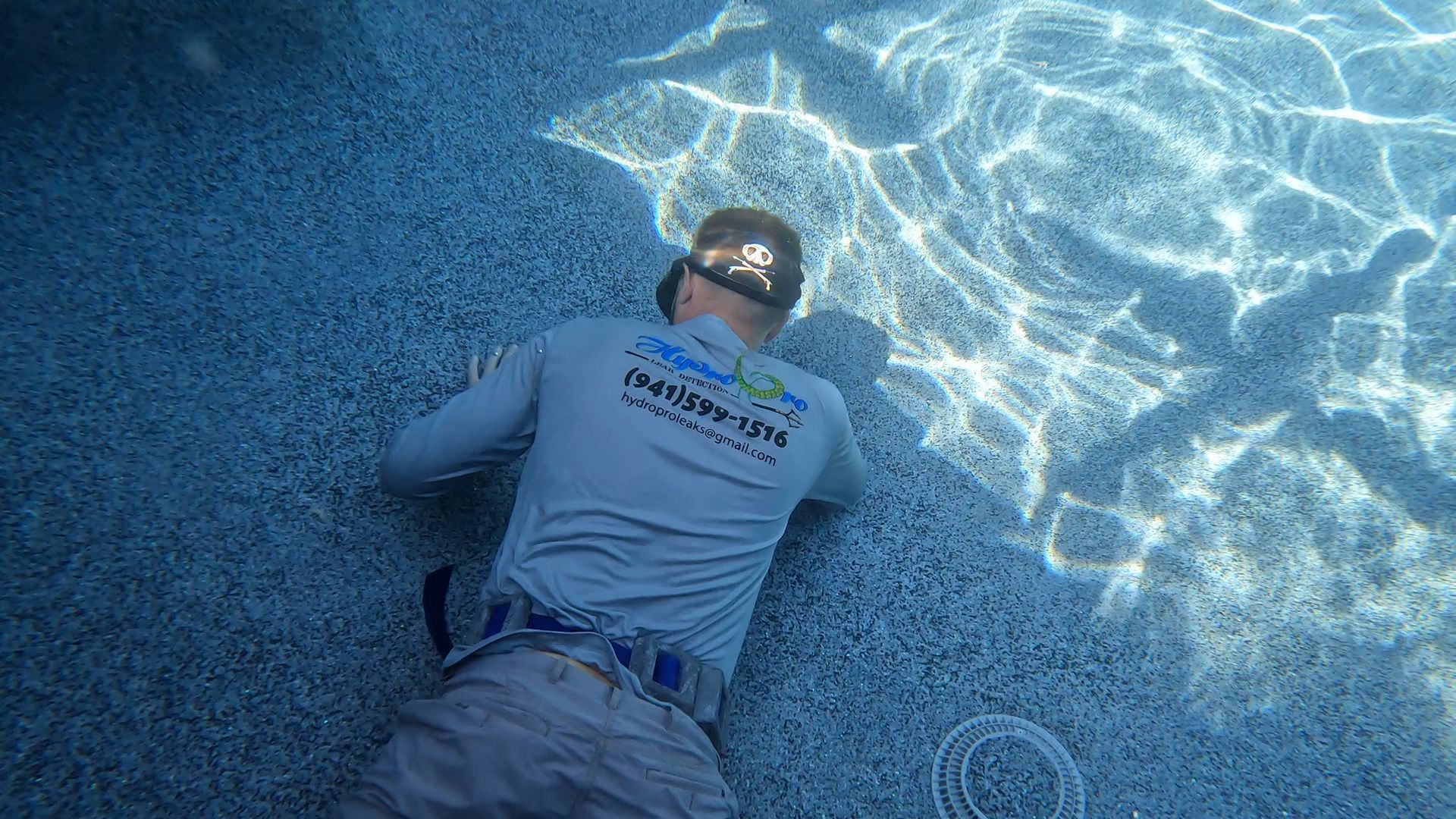 Person floating face-up in a blue pool, wearing a gray shirt and sunglasses, with sunlight rippling on the water