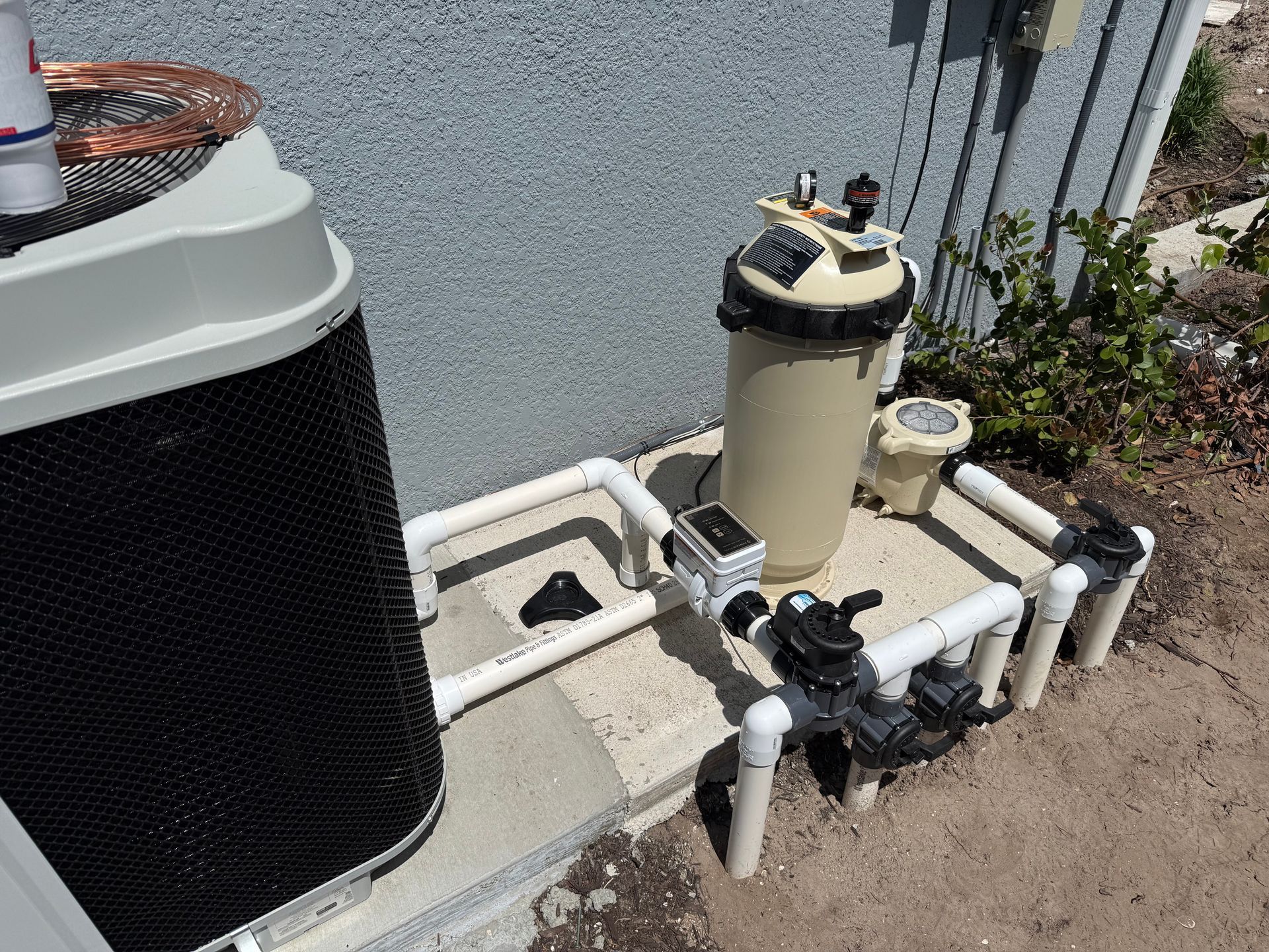 Outdoor pool equipment with pump, filter tank, and white PVC piping beside a house wall.