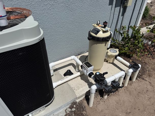 Outdoor pool equipment with pump, filter tank, and white PVC piping beside a house wall.