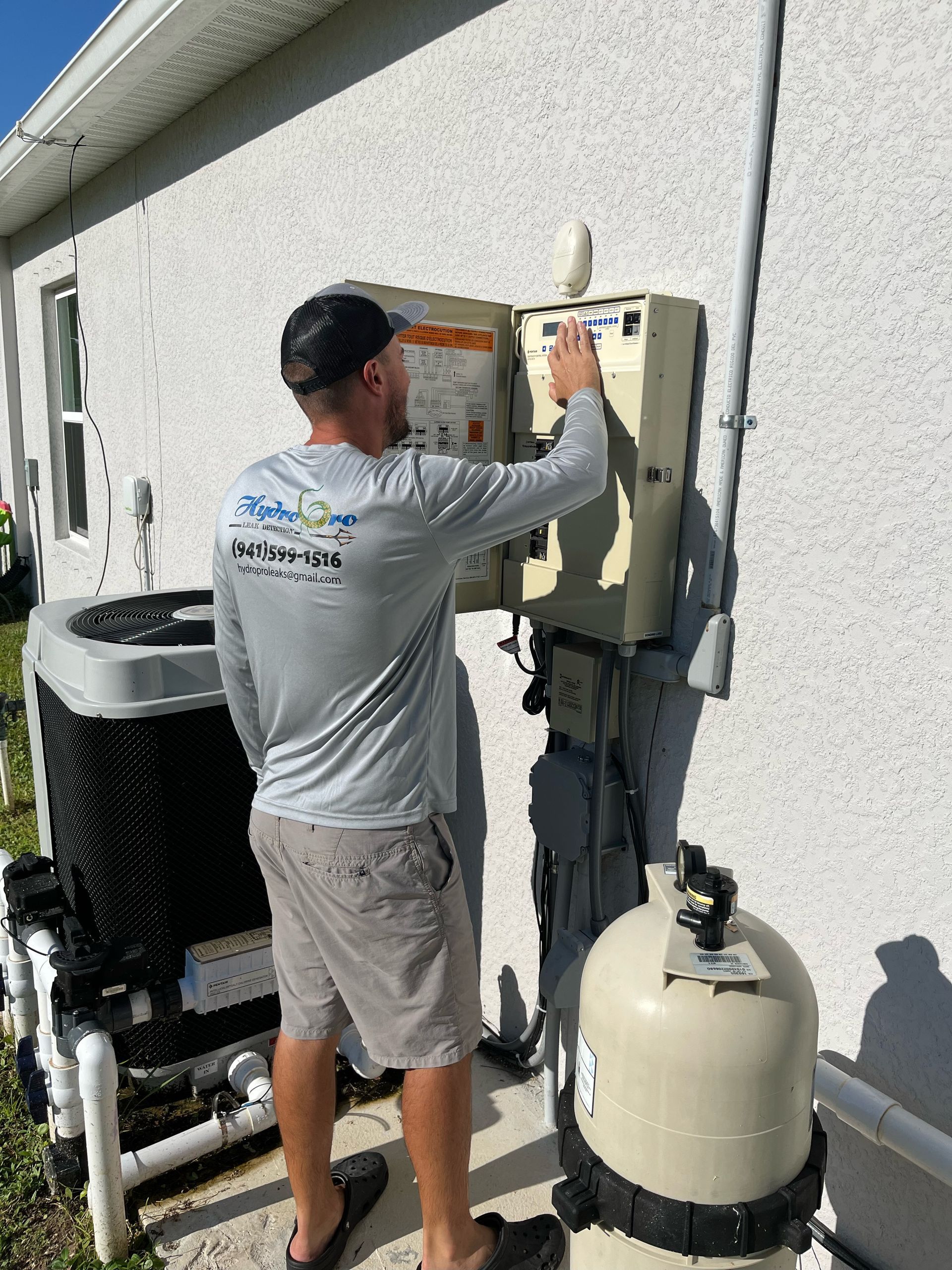 Technician working on outdoor electrical panels beside a pool pump and filter on a house wall