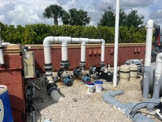 Rooftop plumbing and HVAC equipment with white pipes, valves, and gravel near a red wall.