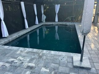 Rectangular backyard pool with dark water, white curtains, and gray stone decking