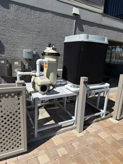Outdoor pool equipment beside a building: black tank, pumps, and white piping on a metal platform.