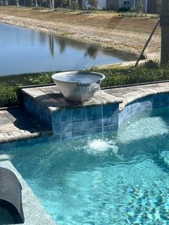 Swimming pool with a stone fountain spilling water, beside a grassy pond and house in the background