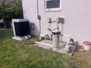 Outdoor utility equipment beside a house wall, including an air conditioner and pool filter unit on grass.