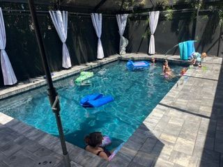 Covered backyard pool with three children and floaties, curtains along the sides and stone deck around the water.