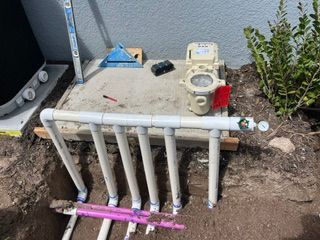 White utility pipes and a water meter in a dirt trench beside a building, with plants nearby