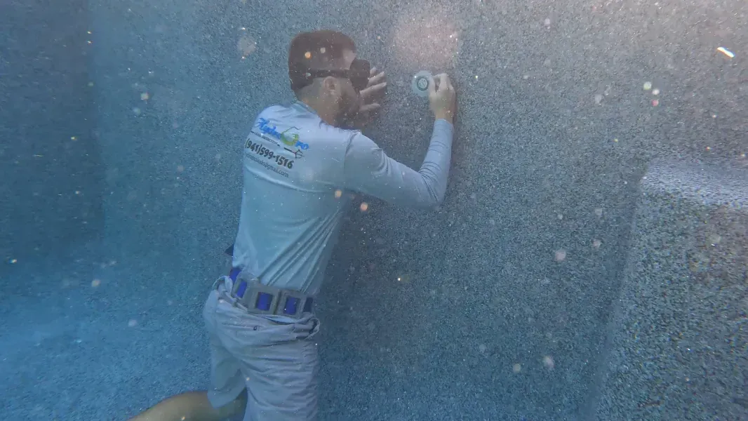 A worker in gray uniform scrapes or inspects a blue wall, surrounded by bubbles or spray underwater.