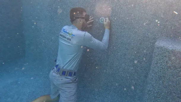 A worker in gray uniform scrapes or inspects a blue wall, surrounded by bubbles or spray underwater.