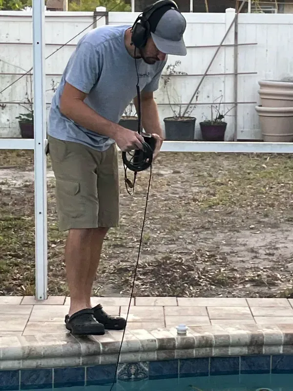 Person using a metal detector beside a backyard pool, leaning over the water with headphones on.