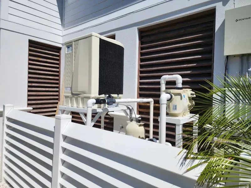 Outdoor HVAC units beside a white fence and house wall, with dark louvered vents and palm fronds nearby