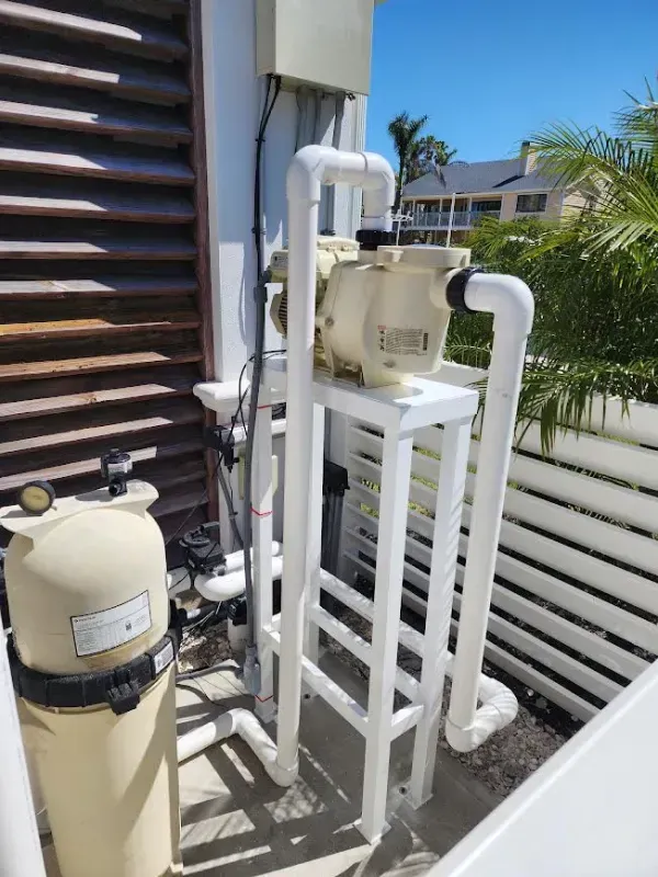 Outdoor pool filtration equipment with white pipes and beige tanks beside a building and fence