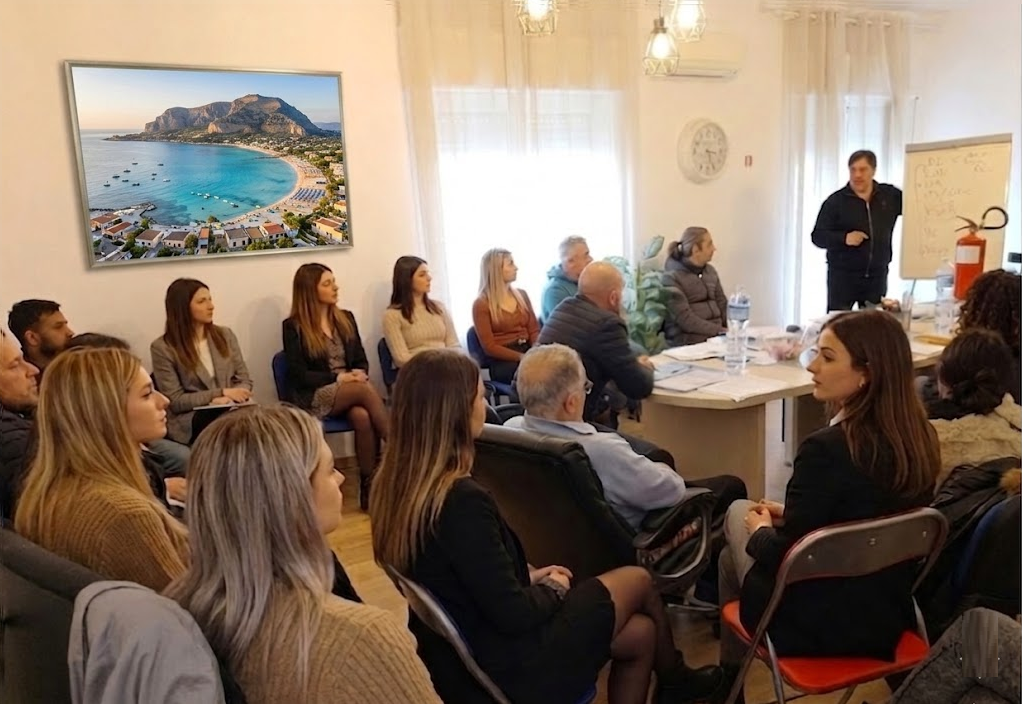 Una piccola sala riunioni con persone sedute attorno a un tavolo, che ascoltano un oratore vicino a una lavagna.