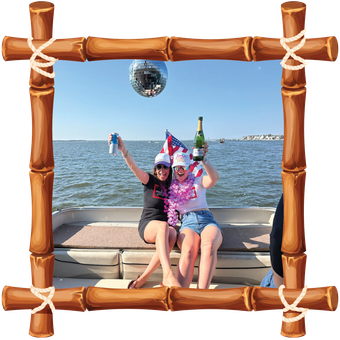 Two people in party hats pose by the sea, framed by a bamboo border, holding up a bottle and drinks.