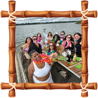 Group of friends on a boat deck, smiling with tropical drinks, framed by bamboo over a waterfront view