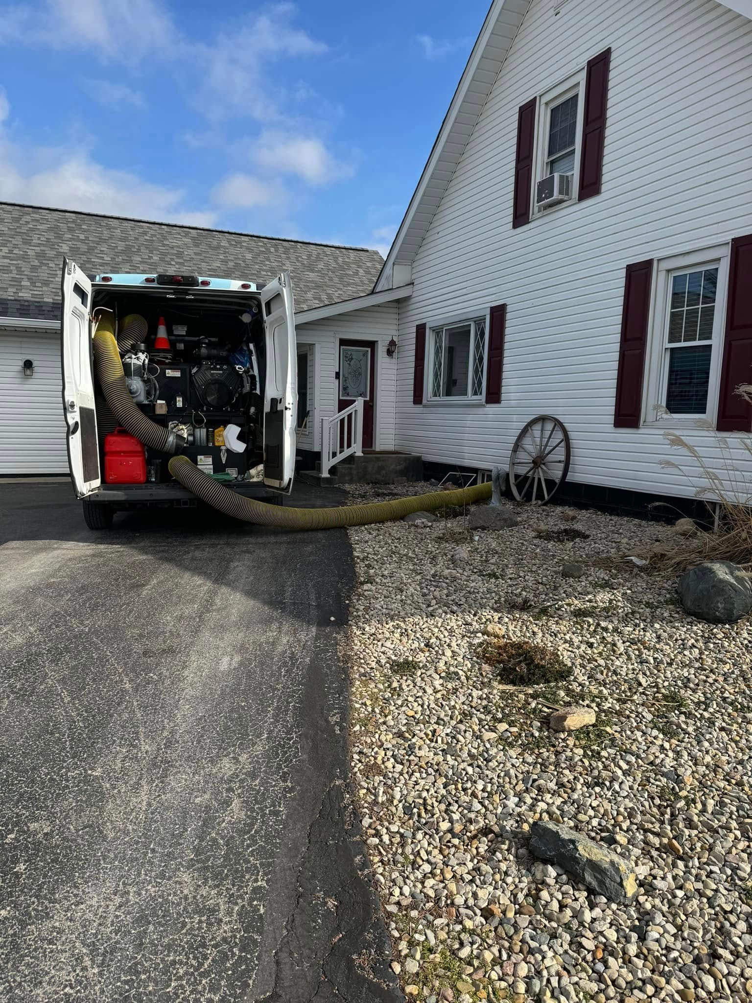 Workers clearing flooded water from a driveway beside a white house with a utility van and hose pump