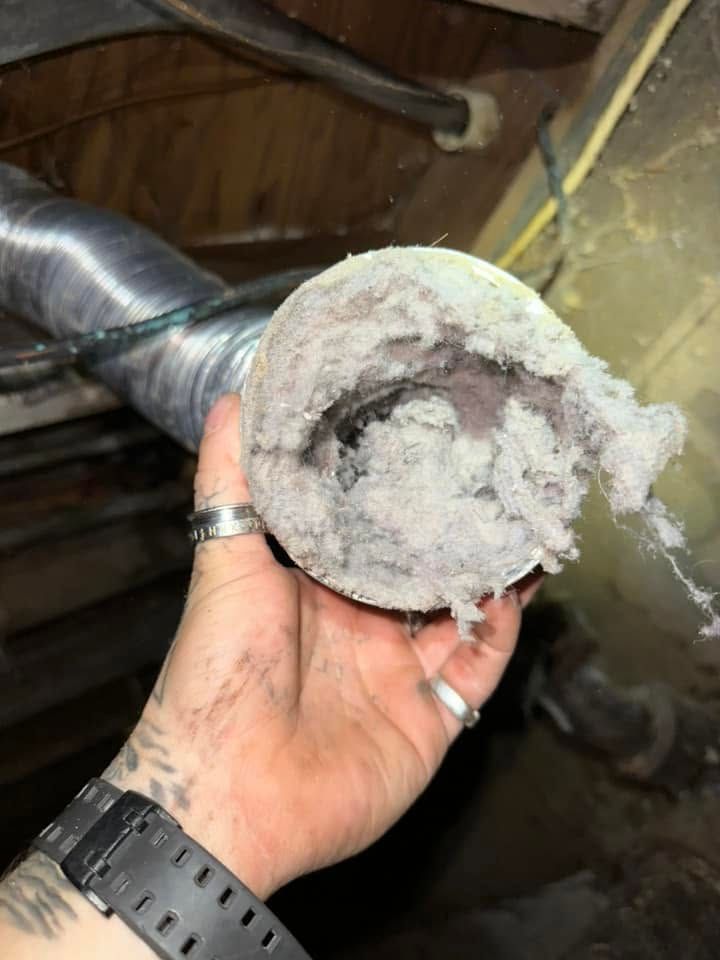 Hand holding a dusty, clogged round air vent or duct opening with insulation debris inside