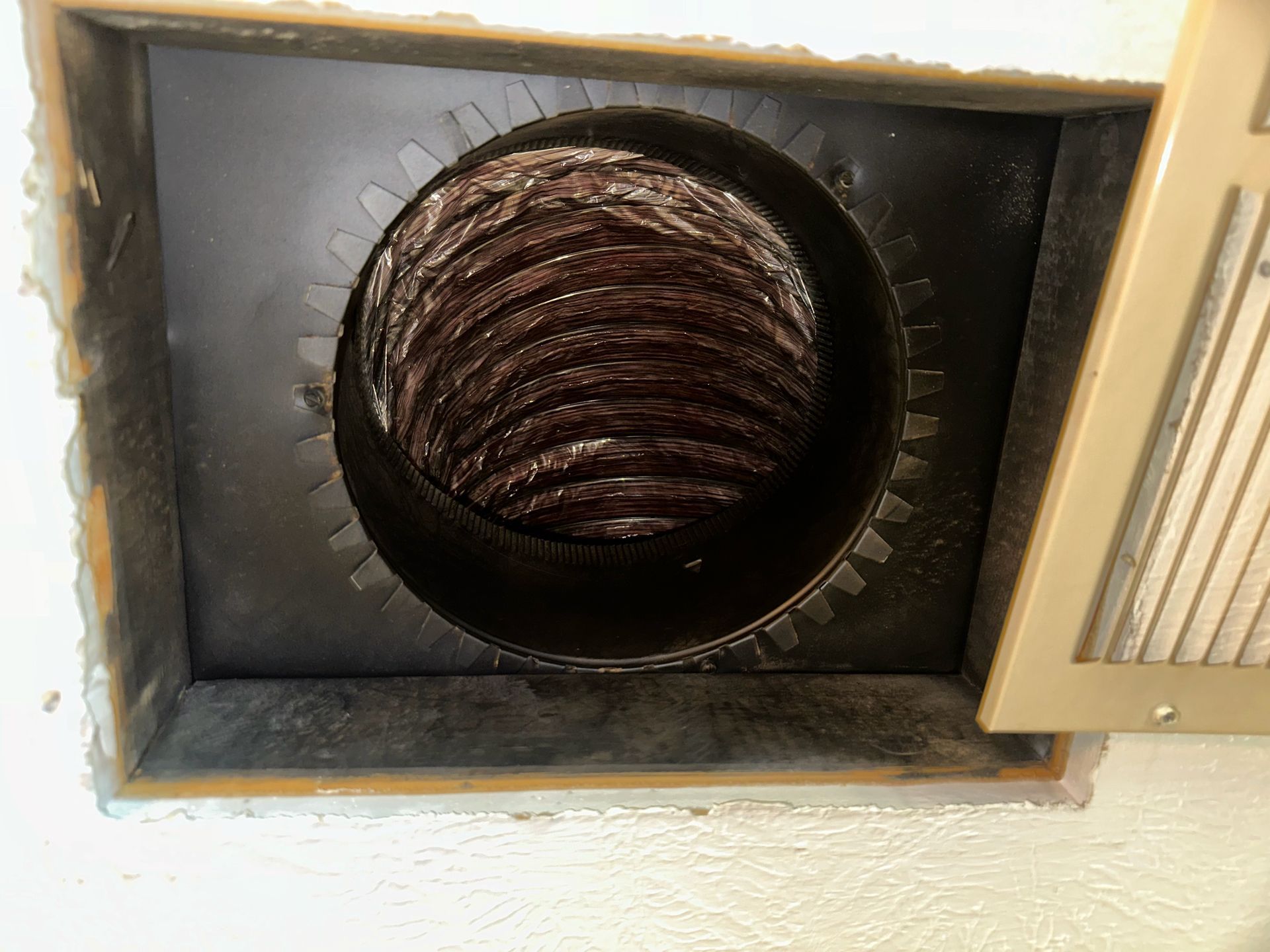 Round black ventilation duct opening in a beige wall with a ribbed hose inside.