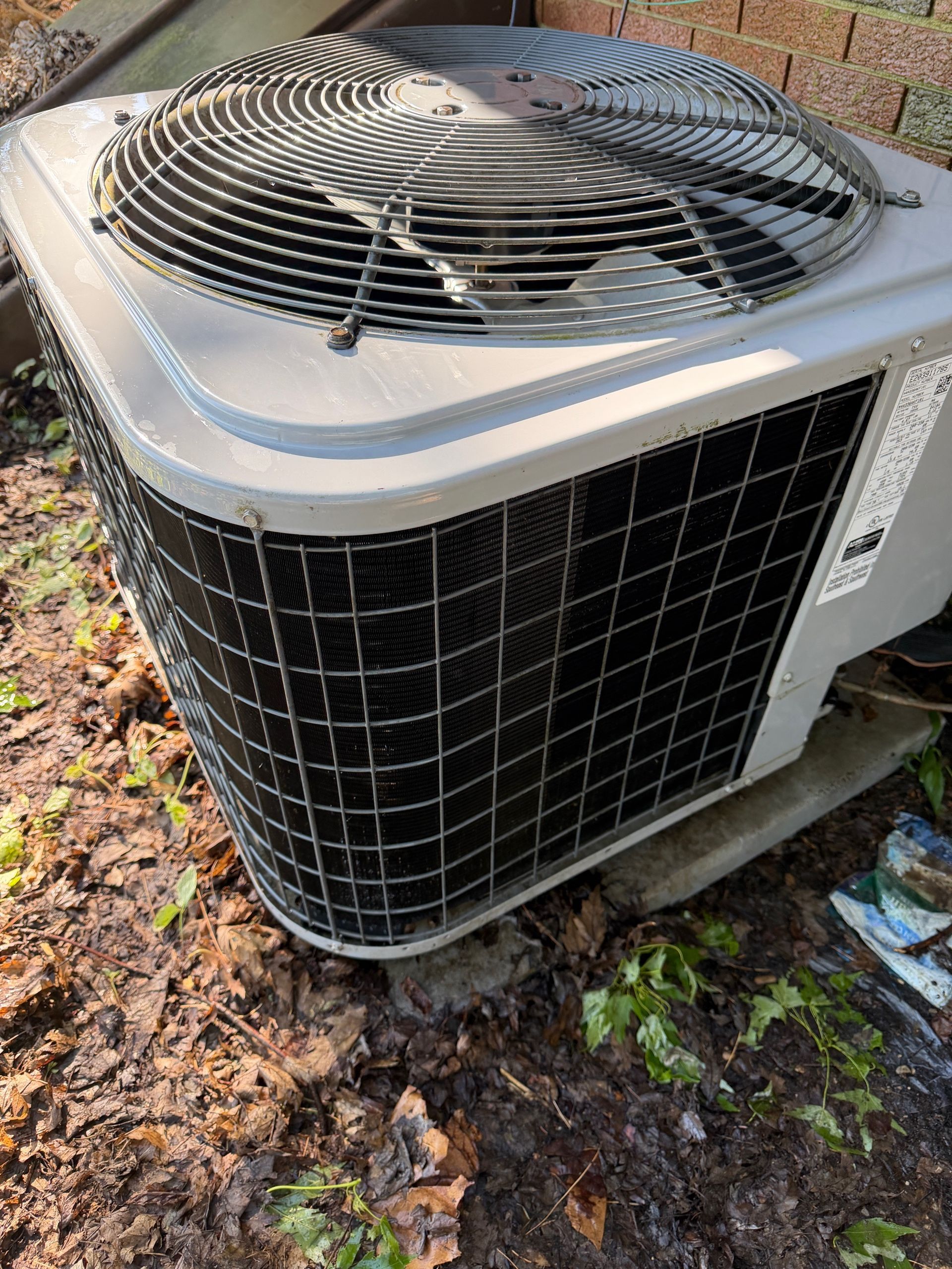 Outdoor air conditioner condenser unit on the ground with a black grille and fan on top, beside leaves and grass